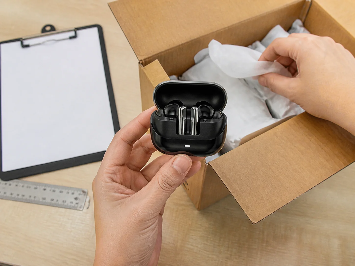 Illustrative receiving scene of the black P103 wireless earbuds being inspected beside an open plain shipping carton and blank clipboard